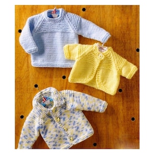 May include: Three knitted baby sweaters, one blue, one yellow, and one white and blue with a speckled pattern. The sweaters are hanging on a wooden surface.
