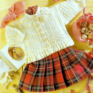 Könnte beinhalten: Ein weißer Strickcardigan mit Rüschenkragen und herzförmigen Knöpfen. Der Cardigan wird mit einem rot karierten Rock kombiniert.