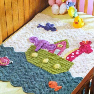 May include: A crocheted baby blanket with a blue, green, and white chevron pattern. The blanket features a crocheted ark with a pink giraffe, purple elephant, and a small pink house. There are also crocheted fish, a turtle, and a bird on the blanket.