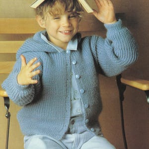 May include: A pale blue knitted cardigan with a hood and button fastening. The cardigan is worn over a light blue collared shirt. The buttons on the cardigan are also pale blue. The cardigan is made of a soft, textured yarn.