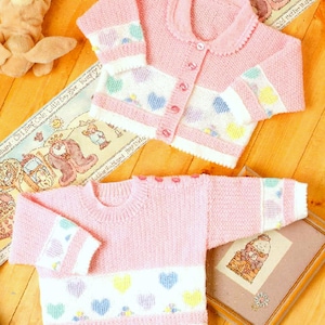 Peut inclure: Deux pulls en tricot rose pour bébé avec des bordures blanches et des motifs de cœurs colorés. Les pulls sont disposés sur une surface en bois.