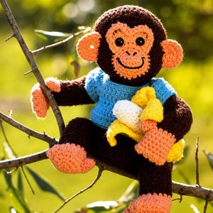 Pode incluir: Um brinquedo de macaco em crochê com pelo castanho escuro, rosto, orelhas, mãos e pés em laranja claro. O macaco veste uma camisola azul brilhante em crochê e segura uma banana em crochê amarela e branca. Olhos pretos e sorriso bordado.