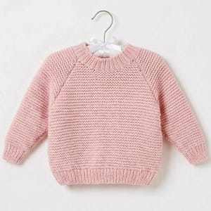 May include: A light pink long-sleeve jumper featuring a textured garter stitch knit pattern, a ribbed crew neckline, ribbed cuffs, and a ribbed hem. The jumper is displayed on a silver metal hanger with a white satin ribbon bow at the top.