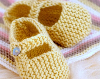 May include: Two pairs of knitted baby shoes in a pale yellow color. The shoes have a simple design with a strap across the top and a button detail. The shoes are sitting on a striped blanket.