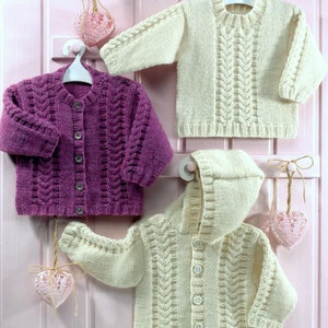 May include: Three knitted baby sweaters in cream and purple hang on a pink wall. The sweaters include a hooded cardigan, a button-up cardigan, and a crew neck sweater. Each sweater has a cable knit design.