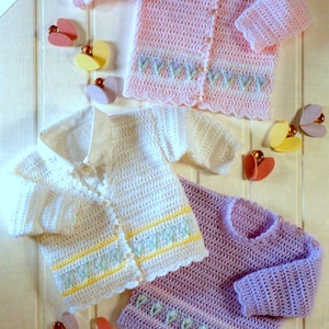 May include: Three crocheted baby cardigans in pink, white, and purple with a scalloped edge and embroidered flower details.  The cardigans are laid out on a light wood surface.
