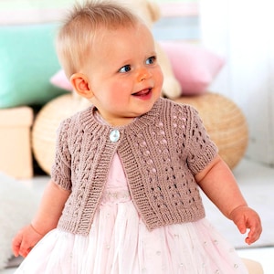 May include: A light brown, short-sleeved knitted cardigan with a decorative button. The cardigan has an openwork design and is worn over a pink dress. The baby is smiling.