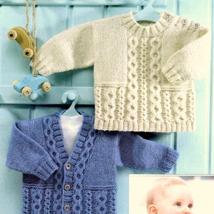 May include: Two knitted baby sweaters, one white and one blue, with a cable knit pattern. The sweaters are hanging on hangers against a light blue wooden background.