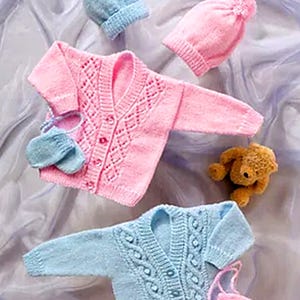 Könnte beinhalten: Zwei gestrickte Baby-Cardigans, einer rosa und einer blau, mit passenden Mützen und Fäustlingen. Die Cardigans haben unterschiedliche Strickmuster und Knopfverschlüsse.