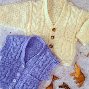 May include: A knitted baby cardigan and vest set in yellow and purple with a cable knit pattern. The cardigan has long sleeves and buttons down the front. The vest is sleeveless and has buttons down the front. Both pieces have pockets.