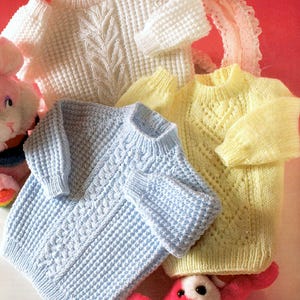 Puede incluir: Tres suéteres de bebé tejidos en crema, azul claro y amarillo. El suéter crema tiene un diseño de hojas, el azul tiene un diseño de punto de cable y el amarillo tiene un diseño de diamantes. Los suéteres están sobre un fondo rojo.