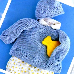 Op de afbeelding: Een lichtblauwe gebreide babyoutfit, bestaande uit een trui, een muts en een broekje met bloemenprint. De trui heeft een witte kraag en bladvormige details. Een gele houten beer zit in de trui.