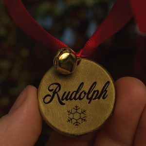 May include: A gold-colored Christmas ornament with the name "Rudolph" in elegant script and a snowflake design. The ornament is attached to a red ribbon and has a small gold bell.
