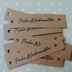 Könnte beinhalten: Ein Stapel rechteckiger brauner Papier-Geschenkanhänger. Jeder Anhänger hat oben ein kleines Loch und trägt den Schriftzug "Frohe Weihnachten" in schwarzer Schrift sowie einen schwarzen Stern. Die Anhänger liegen auf einer hellgrauen Oberfläche mit Schneeflockenmuster.