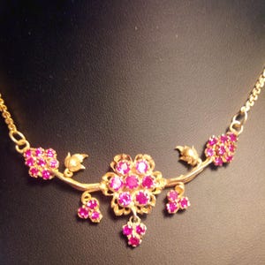 Könnte beinhalten: Eine goldene Halskette mit einem floralen Design, das mit rosafarbenen Edelsteinen besetzt ist. Die Halskette hat eine zarte Kette und einen Anhänger mit einem Cluster aus rosafarbenen Steinen.