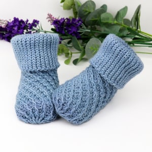 May include: A pair of blue knitted baby booties with a cable knit pattern. The booties have a folded cuff and are made with a soft yarn.