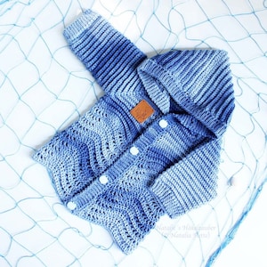 Könnte beinhalten: Ein blauer und weißer gehäkelter Baby-Pullover mit Kapuze und Knopfleiste. Der Pullover hat ein Wellenmuster und ist auf einem blauen Fischernetz fotografiert.