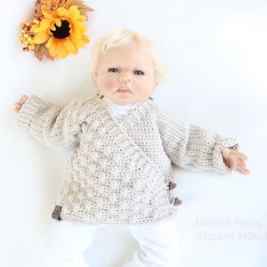 May include: A baby wearing a beige crocheted sweater with a wrap style and brown buttons. The sweater has a textured, bobbly design.