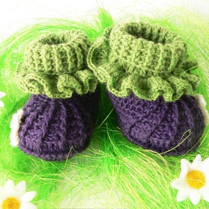 May include: A pair of crocheted baby booties with a purple body and green ruffled trim. The booties are sitting on a bed of green grass.