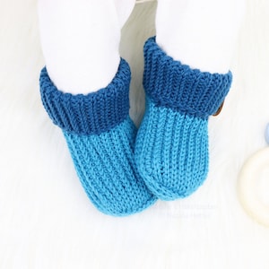 Könnte beinhalten: Ein Paar blaue gestrickte Babyschuhe mit einem dunkelblauen Bündchen. Die Schuhe liegen auf einer weißen Oberfläche.