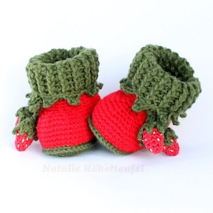 May include: A pair of red and green crocheted baby booties shaped like strawberries. The booties have green leaves and white seeds.