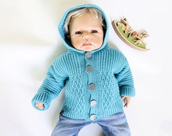 Könnte beinhalten: Ein blauer gestrickter Kapuzenpullover mit Knöpfen. Der Pullover hat ein Rautenmuster und wird von einer Baby-Puppe getragen.