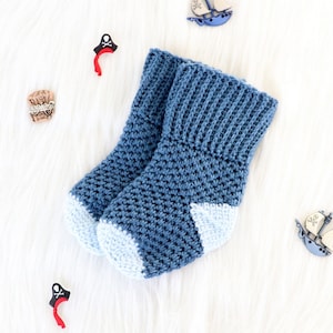 Könnte beinhalten: Ein Paar handgefertigte blaue Baby-Häkelstrümpfe mit hellblauen Zehen und Bündchen. Die Socken liegen auf einer weißen, flauschigen Oberfläche, mit dekorativen Piraten- und Segelboot-Verzierungen.