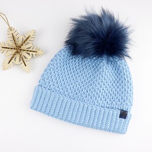 Könnte beinhalten: Eine hellblaue Strickmütze mit einem flauschigen dunkelblauen Bommel oben. Die Mütze hat ein strukturiertes Muster und ein kleines dunkelblaues Etikett an der Seite.