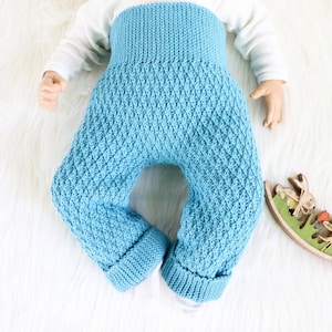 Könnte beinhalten: Eine blaue gestrickte Babyhose mit Rautenmuster. Die Hose ist gefaltet und flach auf einer weißen, flauschigen Oberfläche gelegt.