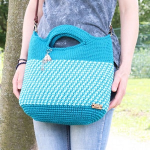 May include: A teal and white crocheted tote bag with a brown leather strap. The bag has a small wooden tag that says "From the Hook".