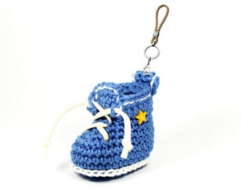 Op de afbeelding: Een blauwe gehaakte babyschoen met witte veters en een gele sterbedel bevestigd aan een bruine koordsleutel.