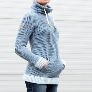 Könnte beinhalten: Ein hellblauer Strickpullover mit hohem Kragen und weißem Saum. Der Pullover hat zwei Taschen und ein kleines Logo-Patch am linken Ärmel.