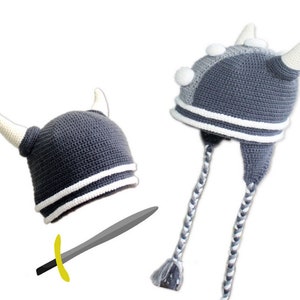 Puede incluir: Gorro de vikingo tejido a ganchillo en tonos grises con detalles blancos, incluyendo cuernos y rayas. El gorro tiene orejeras con lazos trenzados. También se incluye una espada de juguete gris y amarilla.