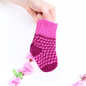 May include: A pair of pink and purple crocheted baby socks. The socks have a textured pattern and are held in a hand against a white background.