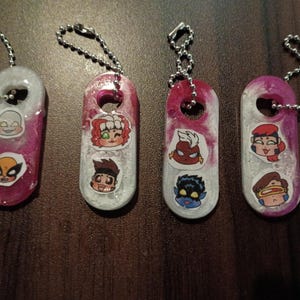 May include: Four oval-shaped resin keychains with silver ball chains. Each keychain features a marbled pink and white finish and two small cartoon character stickers. The characters include various figures with distinct hair colors and facial expressions.