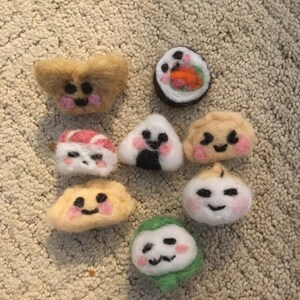 Könnte beinhalten: Eine Sammlung handgefertigter Filz-Lebensmittel, darunter Sushi-Rollen, Onigiri und Knödel. Jedes Stück zeigt lächelnde Gesichter und farbenfrohe Details wie rosa Wangen und Algenakzente. Die Filz-Lebensmittel sind auf einem neutralfarbenen Teppich angeordnet.