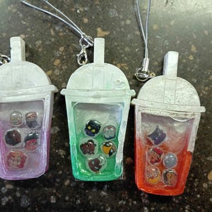 May include: Three bubble tea cup charms with silver lanyards. Each charm features a white lid and a clear body filled with liquid and small circular character icons. The cups have pink, green, and orange gradients at the base respectively.