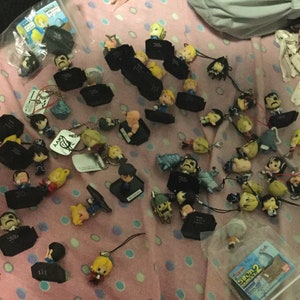 May include: A collection of small character figurines and keychains scattered on a pink patterned surface. The figurines feature various characters with detailed facial features and clothing. Some are attached to black rectangular bases.