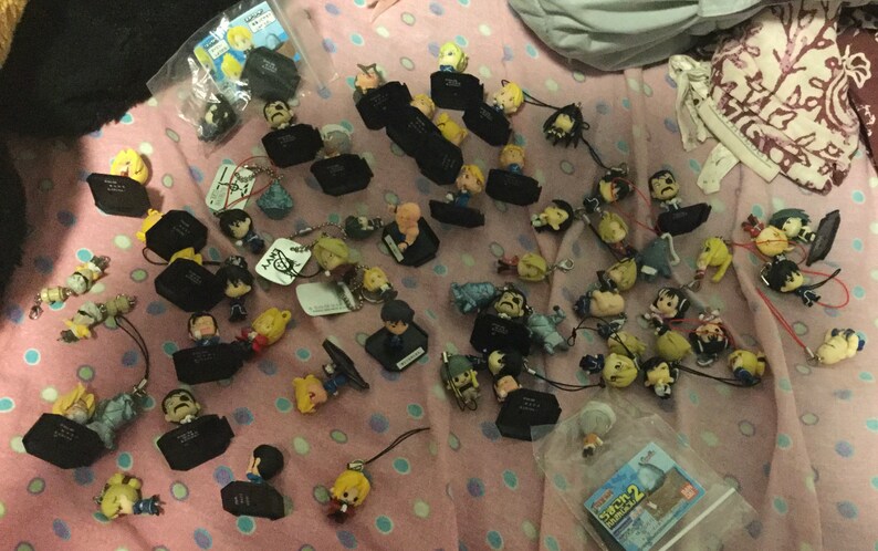 May include: A collection of small character figurines and keychains scattered on a pink patterned surface. The figurines feature various characters with detailed facial features and clothing. Some are attached to black rectangular bases.