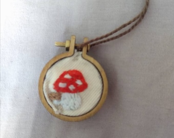 May include: A handmade mushroom pendant necklace. The pendant features a red mushroom with white spots, embroidered on a cream-colored fabric, set in a small wooden hoop. The necklace has a brown cord.