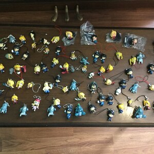 May include: A collection of small anime character figurines with various poses and colors, many with attached lanyards. The figurines are arranged on a dark brown wooden surface.