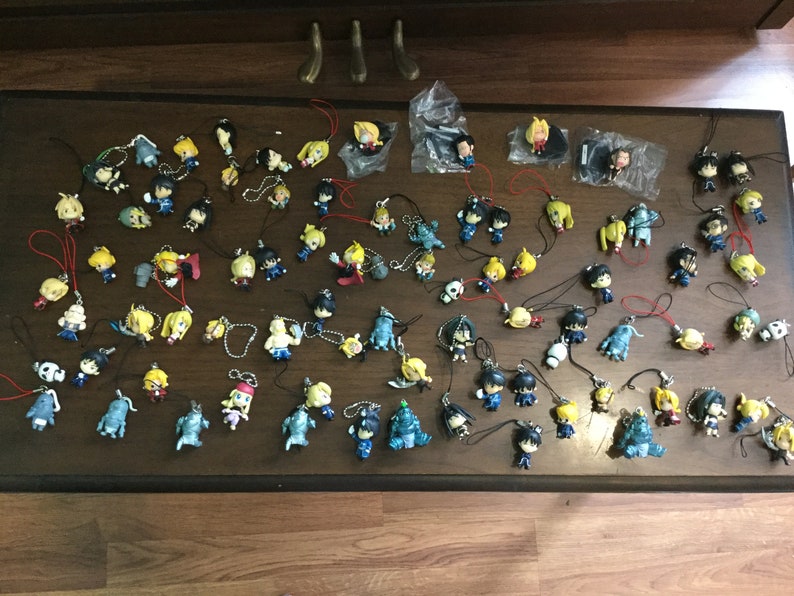May include: A collection of small anime character figurines with various poses and colors, many with attached lanyards. The figurines are arranged on a dark brown wooden surface.