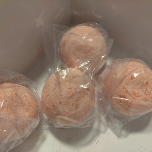May include: Four round, light pink bath bombs in clear plastic packaging are arranged inside a white box. The bath bombs have a textured surface and a speckled appearance, suggesting a blend of ingredients.