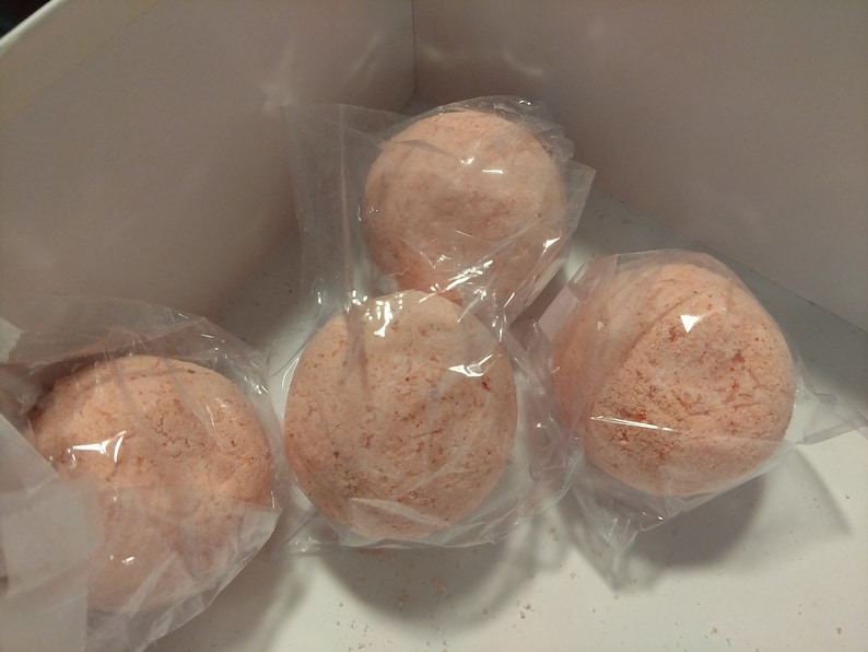 May include: Four round, light pink bath bombs in clear plastic packaging are arranged inside a white box. The bath bombs have a textured surface and a speckled appearance, suggesting a blend of ingredients.