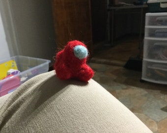 May include: A small, handmade red felted figure with a light blue visor. The figure is a character from the video game Among Us. The plushie is sitting on a tan surface. The figure is a unique, handcrafted item.