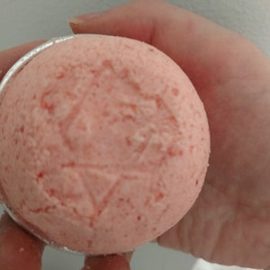 May include: A round, pink bath bomb with a star-like design pressed into the surface. The bath bomb is held in a silver-colored metal container. The bath bomb has a textured appearance with small red flecks.