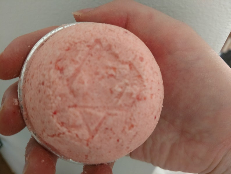 May include: A round, pink bath bomb with a star-like design pressed into the surface. The bath bomb is held in a silver-colored metal container. The bath bomb has a textured appearance with small red flecks.