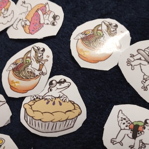 May include: A selection of die-cut stickers showing cartoon frogs in different scenarios. The frogs are illustrated eating ramen, enjoying pie, and interacting with sushi and donuts. The stickers are on a dark blue surface.