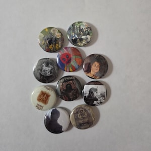 Puede incluir: Una colección de diez botones redondos con varios diseños. Los botones presentan imágenes de obras de arte, retratos y texto. Los colores incluyen negro, blanco, rojo y tonos de marrón. Los botones están dispuestos sobre una superficie blanca.