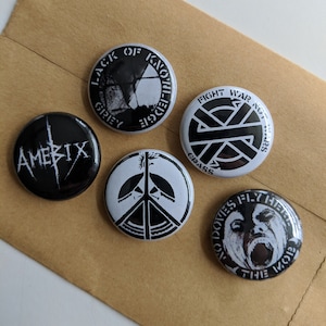 以下が含まれることがあります： 異なるバンドのロゴが描かれた、白黒の丸いボタンが5つ。ロゴは「Lack of Knowledge」、「Amerix」、「Fight War Not Wars」、「Crass」、「No Doves Fly Here The Mob」です。
